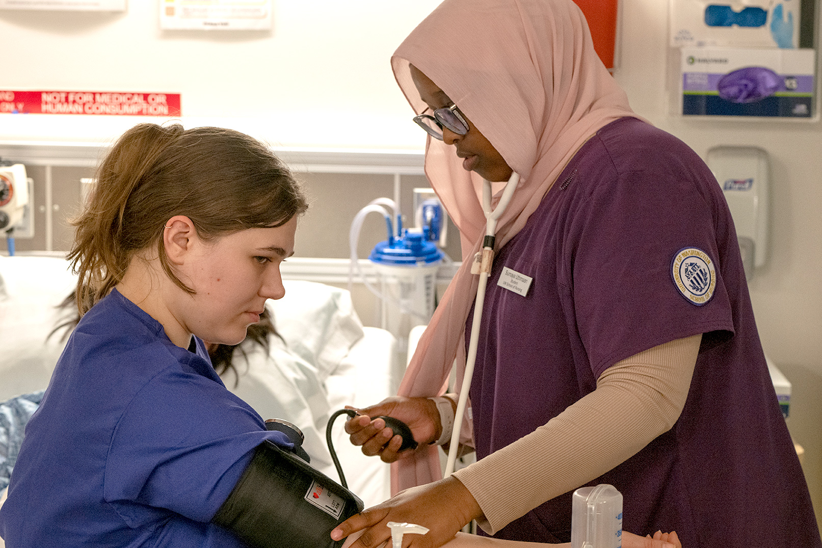 A nursing student in purple scrubs and a hijab takes another student’s blood pressure in a simulation lab